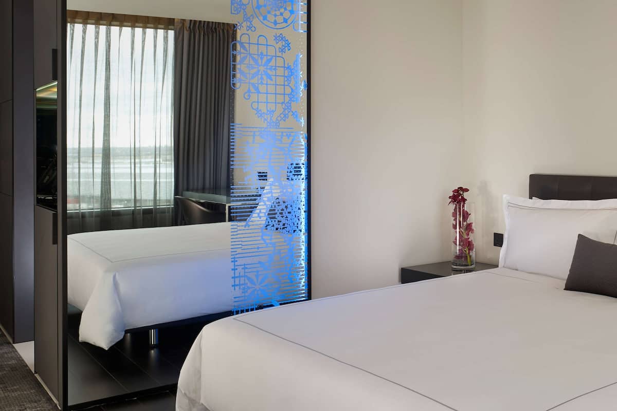 A hotel room with a bed, a small table with a vase, and a view of the sea through sheer curtains.
