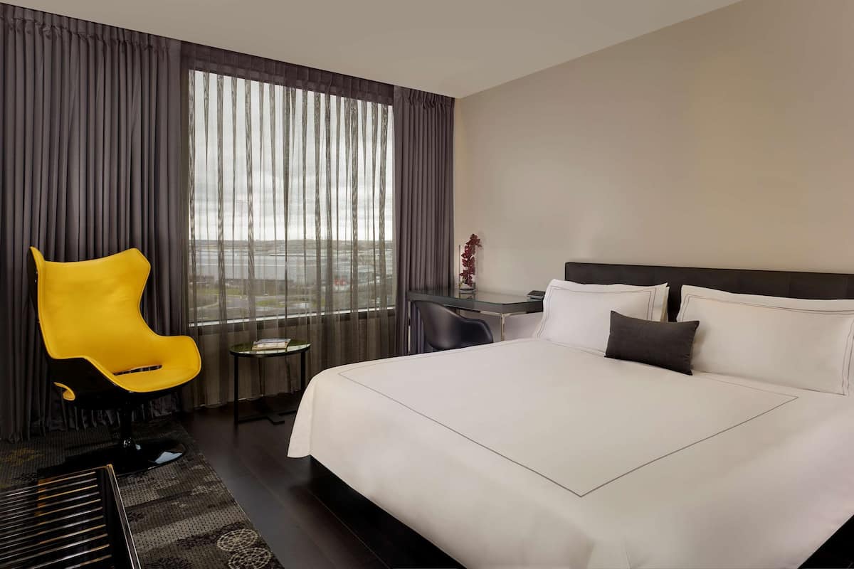 A hotel room with a large bed, a desk, a chair, and a view of the city through sheer curtains.