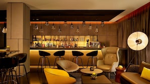 A modern bar area with a bar counter, multiple seating options including bar stools and a sofa, and a variety of bottles and glasses on the shelves. - Park Plaza London Park Royal (London)