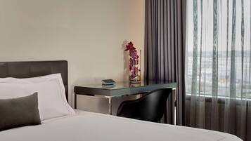 A hotel room with a bed, a small desk with a vase of flowers, a chair, and a view of the city through the window.