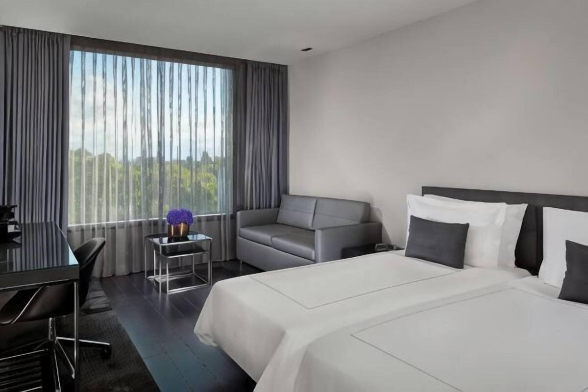 A hotel room with a large bed, a sofa, a desk, and a view of the outdoors through large windows.