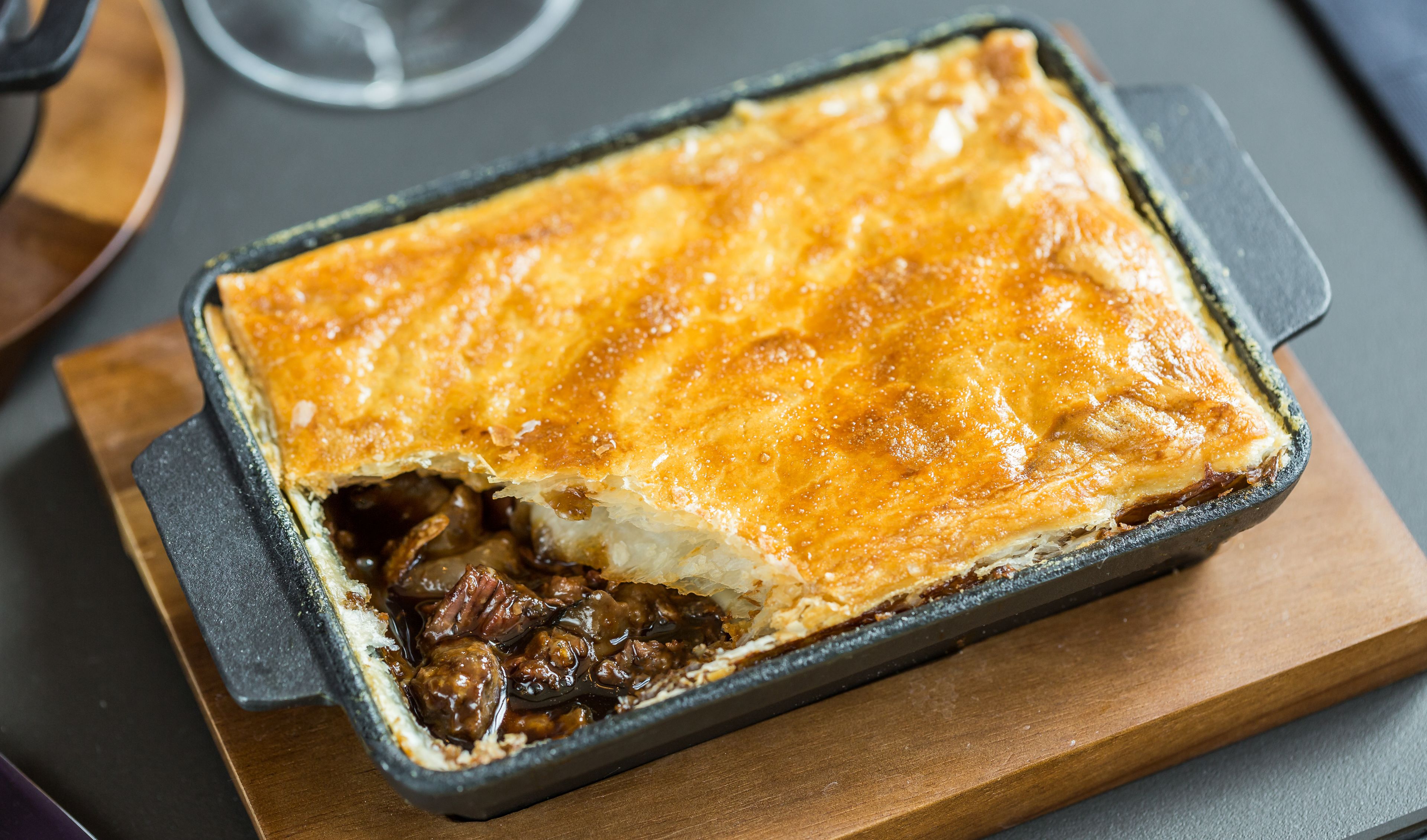 A rectangular baking dish with a golden-brown crust, containing a dark filling, possibly meat, on a wooden board.