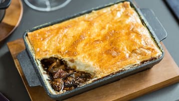 A rectangular baking dish with a golden-brown crust, containing a dark filling, possibly meat, on a wooden board.
