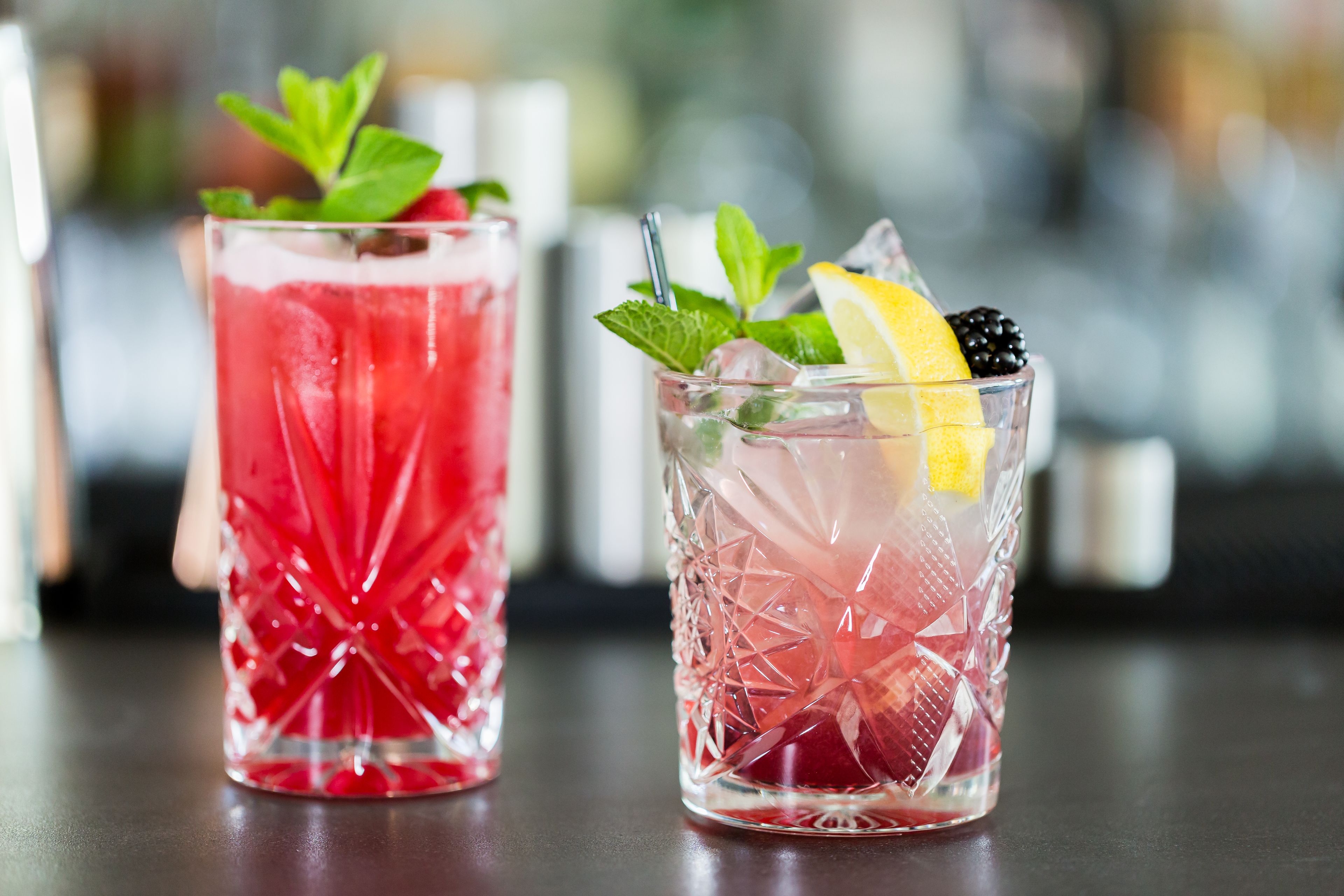 Two glasses of iced drinks with mint leaves and fruit slices on a bar counter.