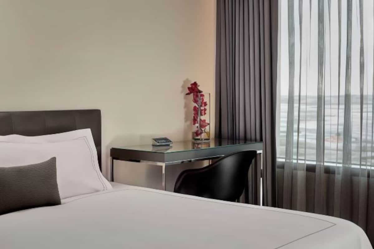Habitación de hotel con cama, un escritorio pequeño, una silla y vistas de la ciudad a través de cortinas translúcidas.