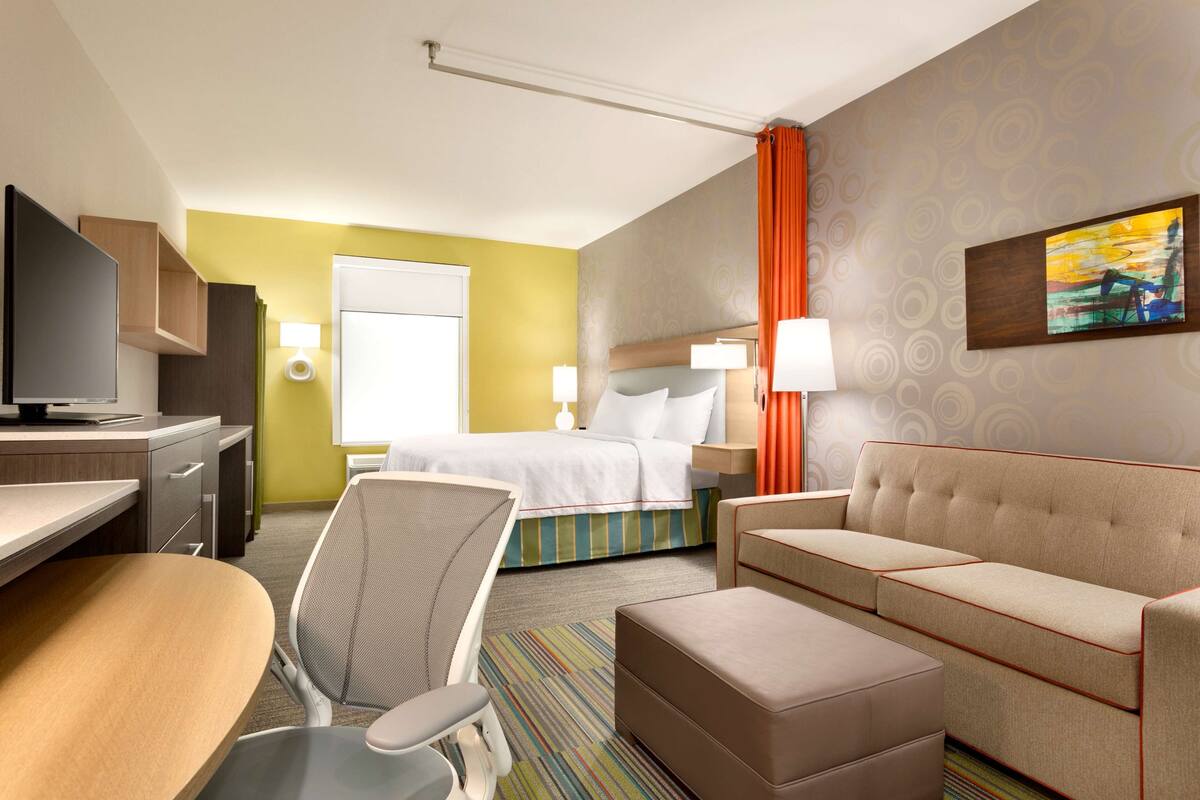 Een hotelkamer met een bed, een slaapbank, een bureau met stoel, een televisie en een raam met gordijnen.