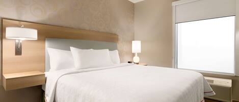 A hotel room with a large bed, two bedside lamps, a headboard, a wall-mounted shelf, and a window with blinds.