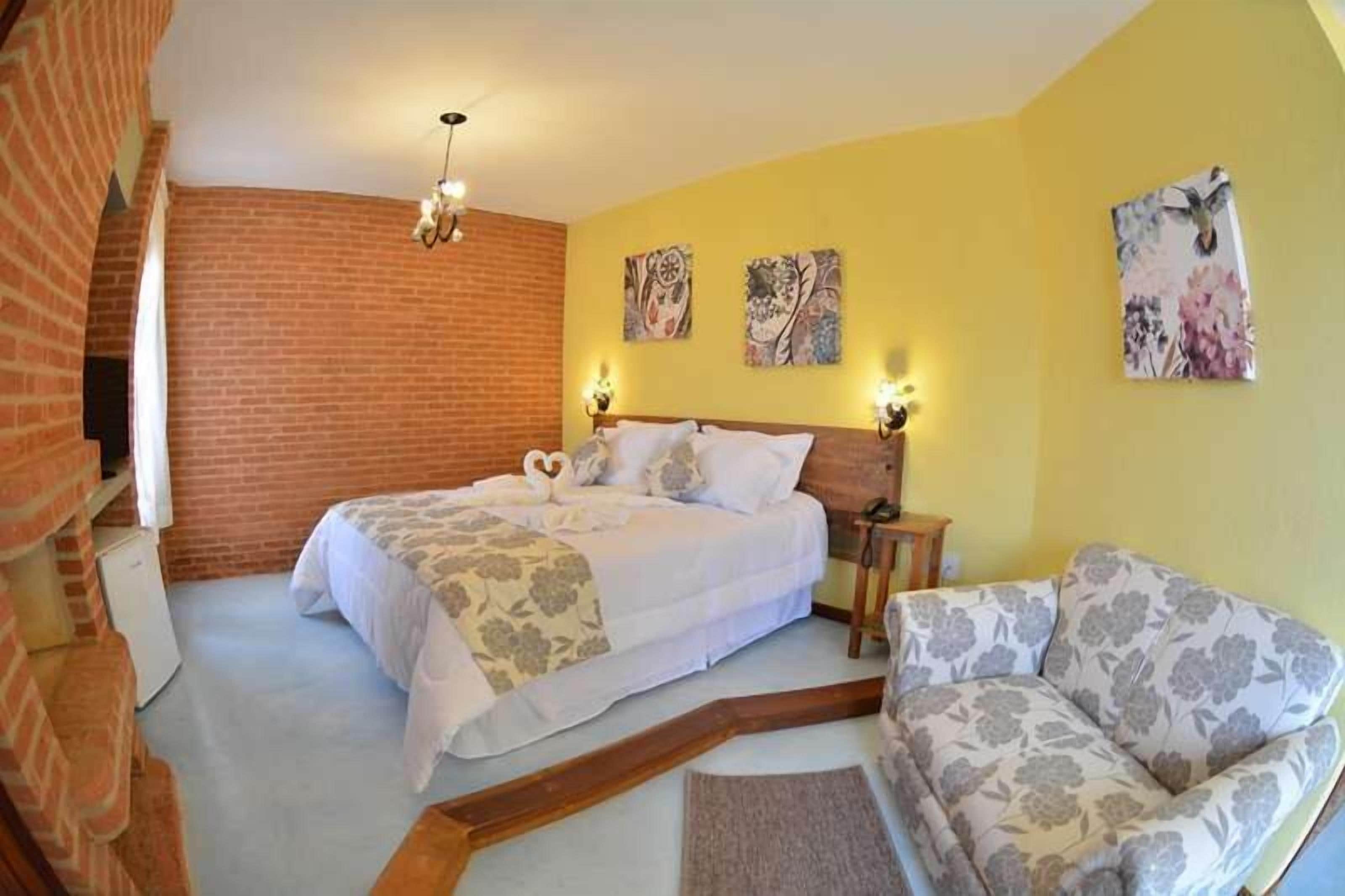 Un dormitorio con una cama, una silla y una mesita. Se observan obras de arte en la pared y una pared de ladrillo.