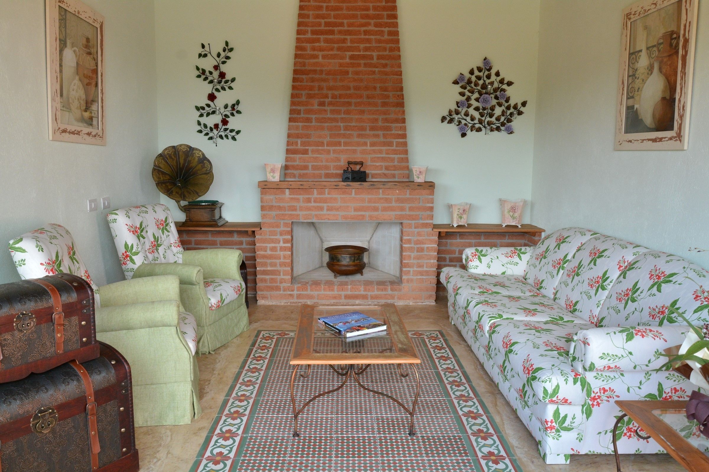 A living room with a brick fireplace, floral-patterned sofas, a green armchair, and a vintage suitcase.