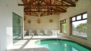 A swimming pool with a curved edge, surrounded by white chairs and tables, under a wooden beam roof, with a brick wall and windows in the background. - Pousada Villa dos Leais (Serra Negra)