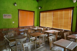 A restaurant with wooden tables and chairs, large windows with blinds, and green walls.