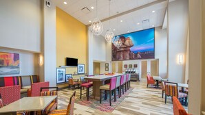 A hotel lobby with a long bar, multiple seating areas, and a large mural on the wall.