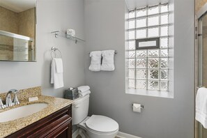 Un baño con lavamanos de encimera de granito, inodoro, cabina de ducha con mampara de vidrio y ventana con paneles de vidrio esmerilado.