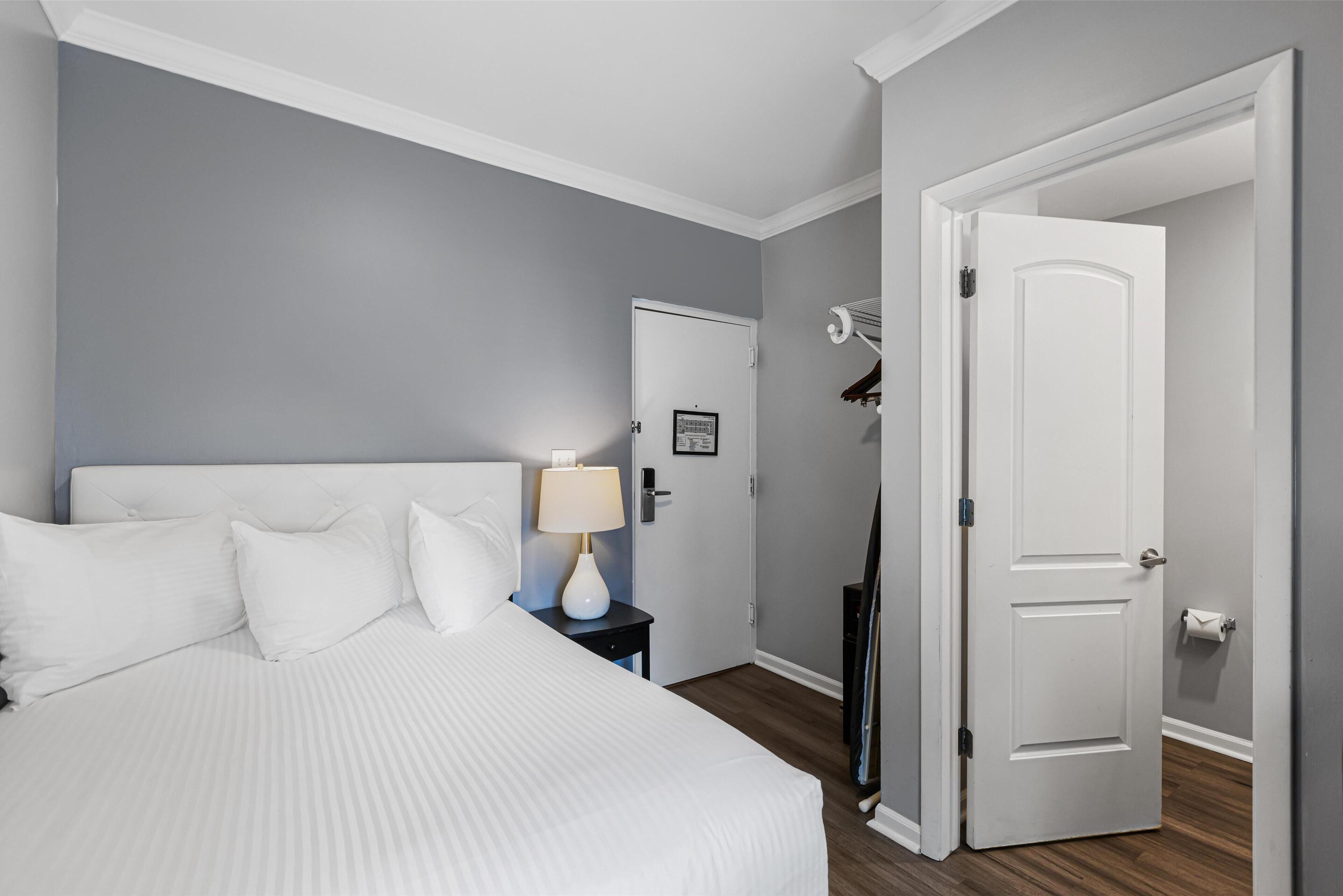 Habitación de hotel con una cama, una mesita de noche con lámpara, una puerta y un perchero.