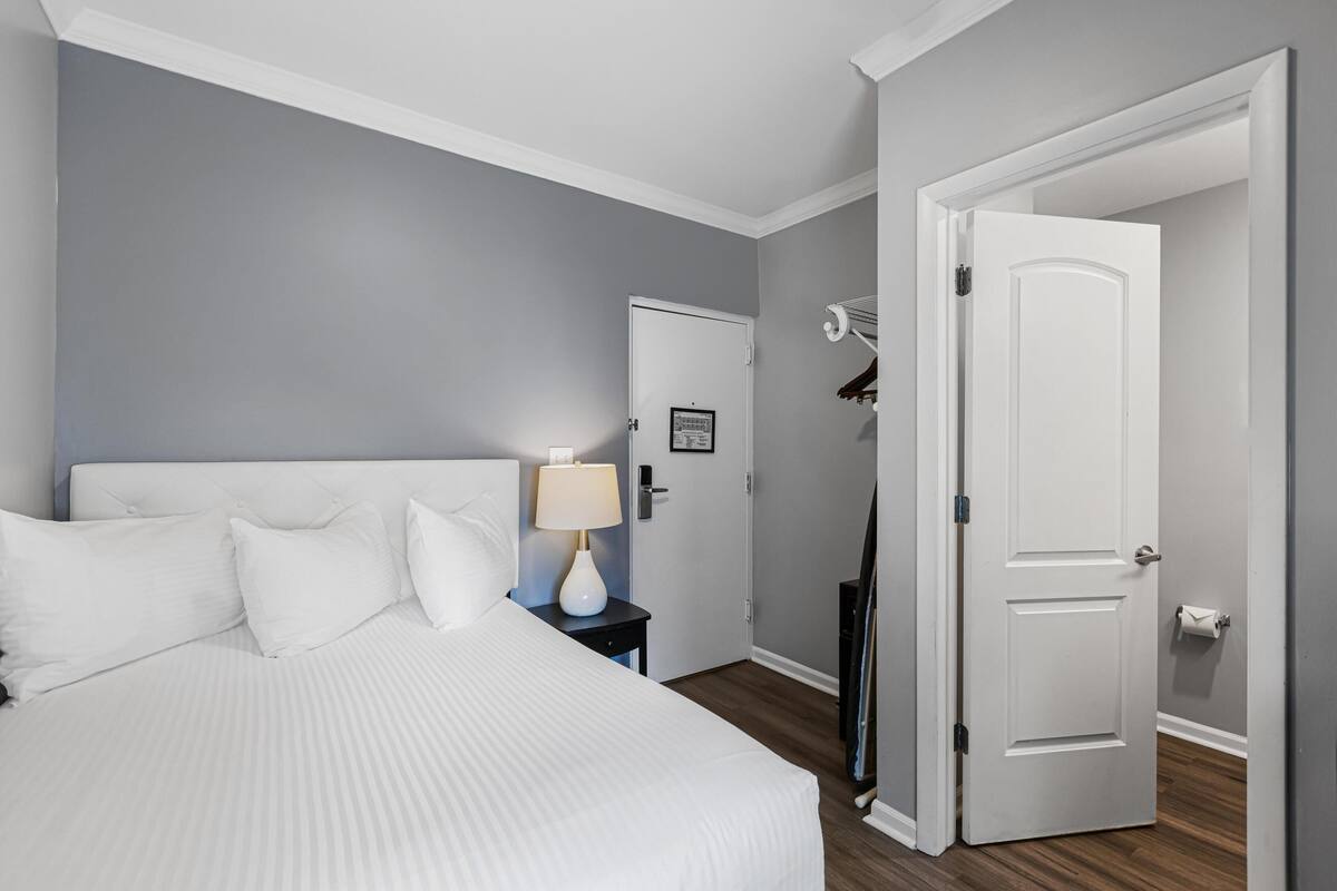 A hotel room with a bed, a nightstand with a lamp, a door, and a coat rack.