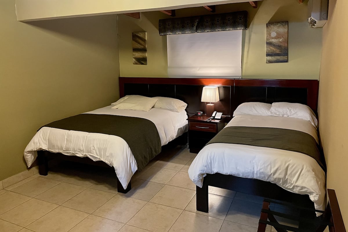 Une chambre d’hôtel avec deux lits simples, une table de chevet avec une lampe, une petite table et une fenêtre avec un rideau.