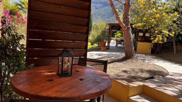 Una pérgola de madera al aire libre con una mesa redonda y sillas, rodeada de plantas en macetas y vistas a una montaña.
