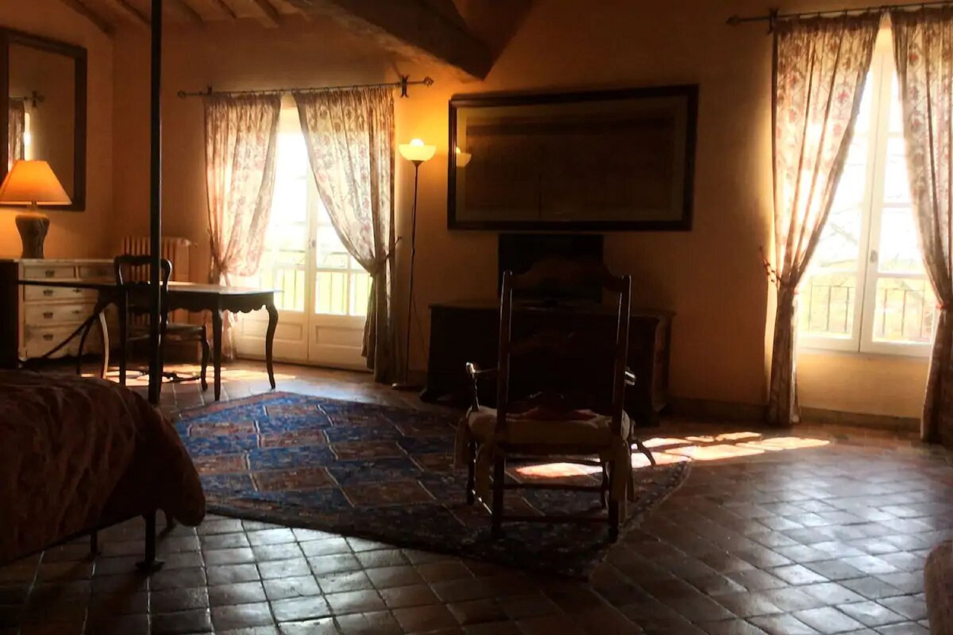 A bedroom with a bed, a chair, a desk, a lamp, a fireplace, and a large window with curtains.