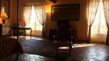 A bedroom with a bed, a chair, a desk, a lamp, a fireplace, and a large window with curtains.