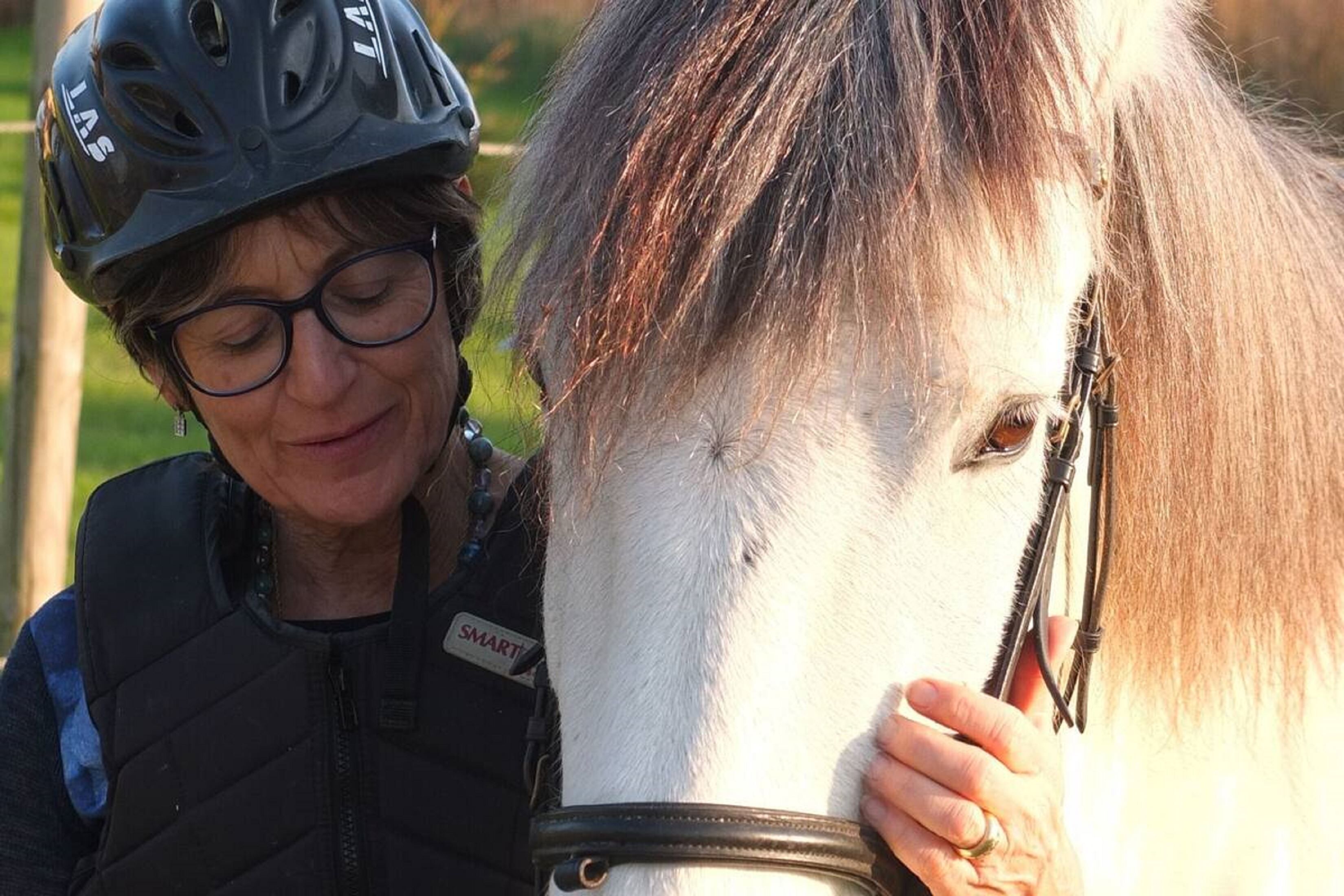 Una persona con casco y chaleco con identificación, acariciando un caballo blanco con brida.