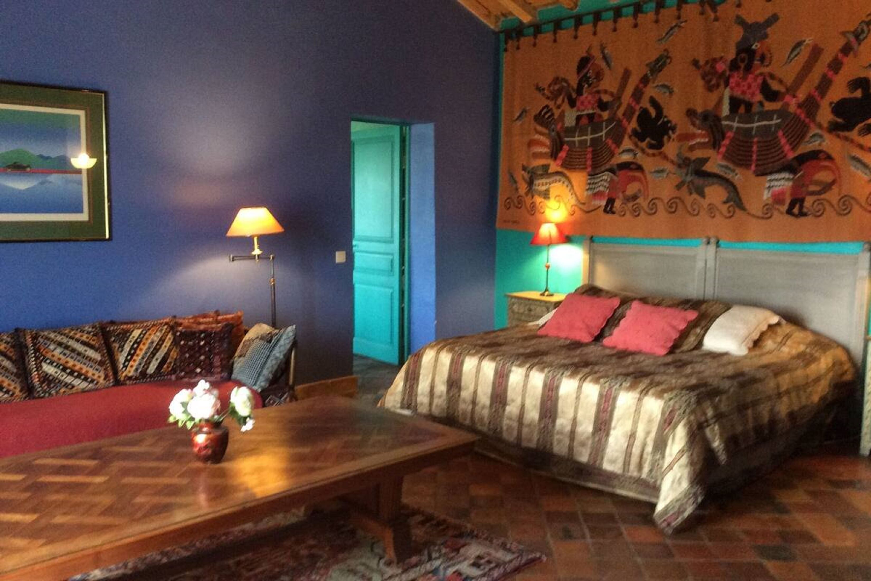 A bedroom with a bed, a sofa, a table with a vase of flowers, a lamp, and a tapestry on the wall.