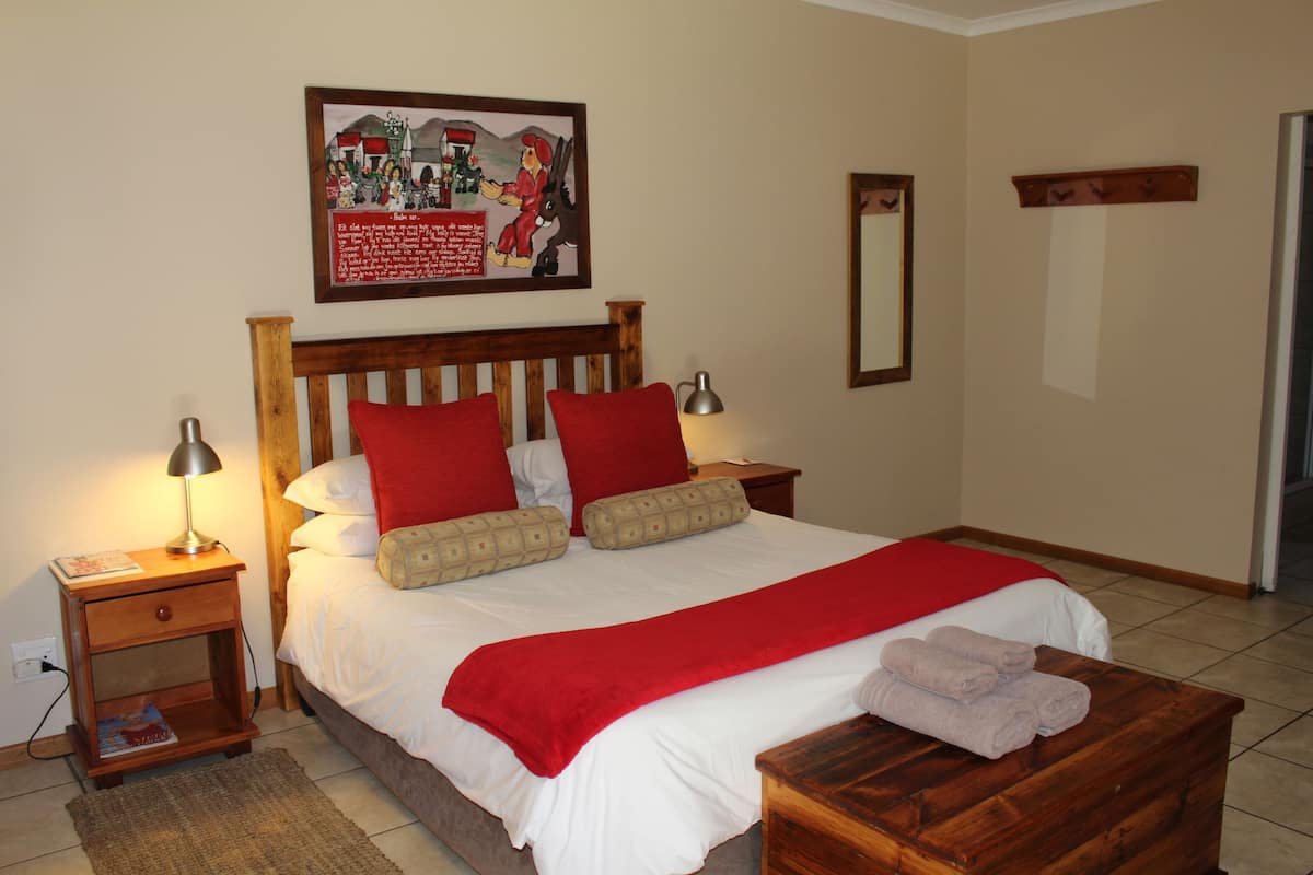 Ein ordentlich bezogenes Bett mit roten und beigen Kissen, einem Holzkopfstück und einer roten Tagesdecke. Ein Nachttisch aus Holz mit einer Lampe, einem Spiegel und einem kleinen Regal mit Büchern steht daneben. Vor dem Bett befindet sich eine Holzbank mit Handtüchern.