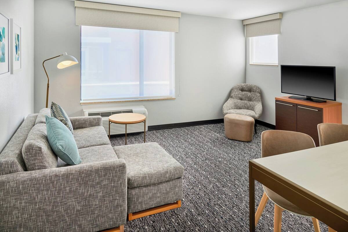 A modern hotel room with a grey sofa, a small round table, a television on a wooden cabinet, and a large window with blinds.