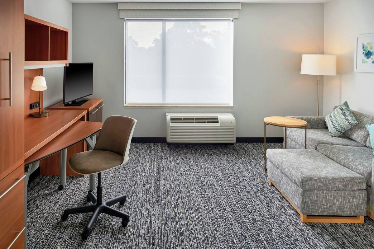 A hotel room with a wooden desk, a television, a grey sofa, a small round table, and a standing lamp.