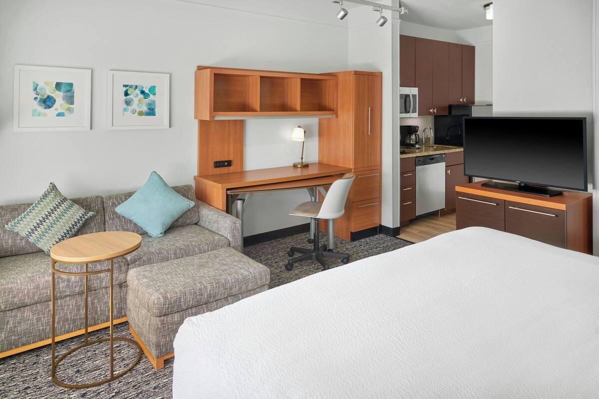 A hotel room with a bed, a sofa, a desk, a chair, a small round table, a television, a kitchen area, and artwork on the walls.