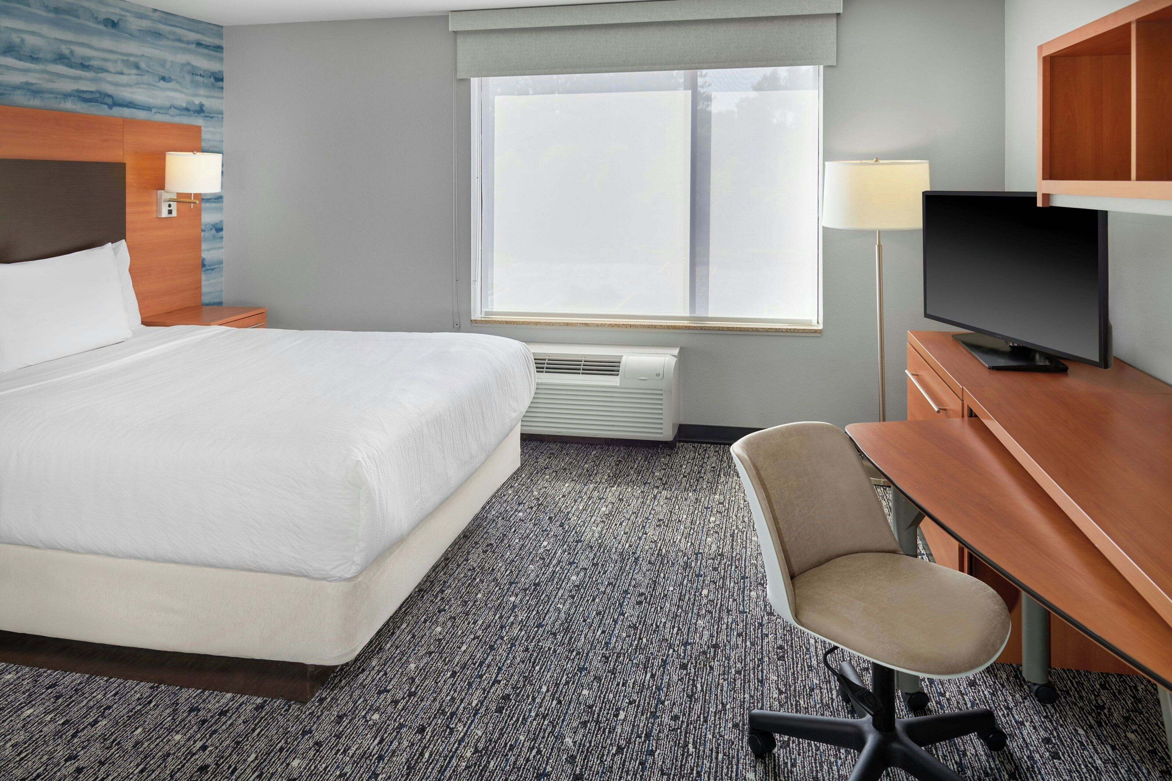 Una habitación de hotel con una cama grande, un escritorio con silla, televisor y ventana con persianas.