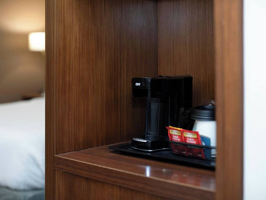 A wooden bedside table with a coffee maker and a cup of coffee, a tray with a packet of sugar and a small container.