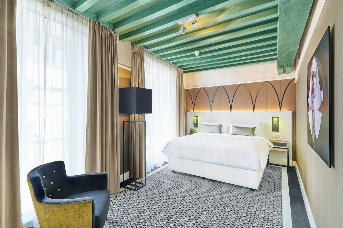 A hotel room with a large bed, a desk, a chair, a standing lamp, and a large window with curtains.