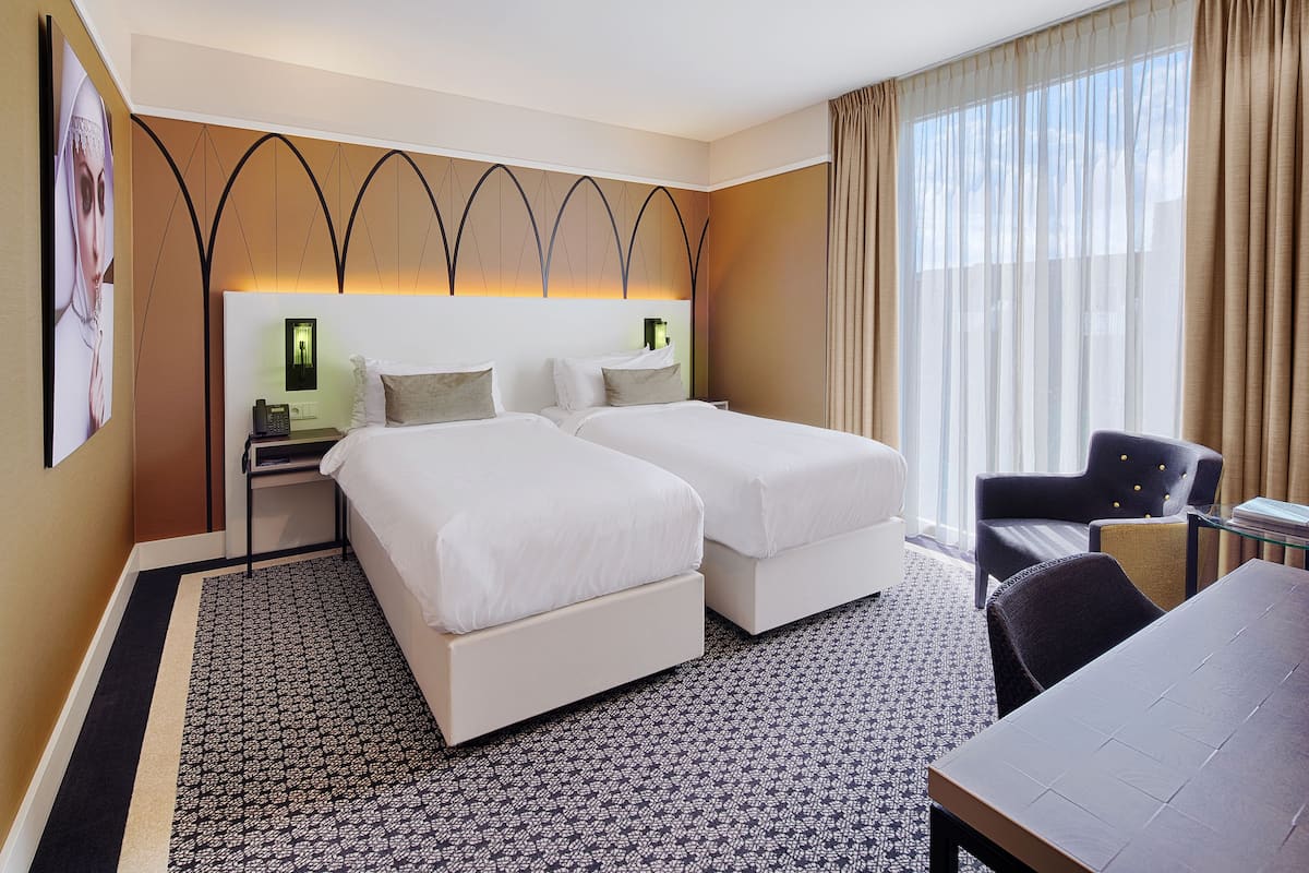 A hotel room with two beds, a desk, a chair, and a large window with curtains.