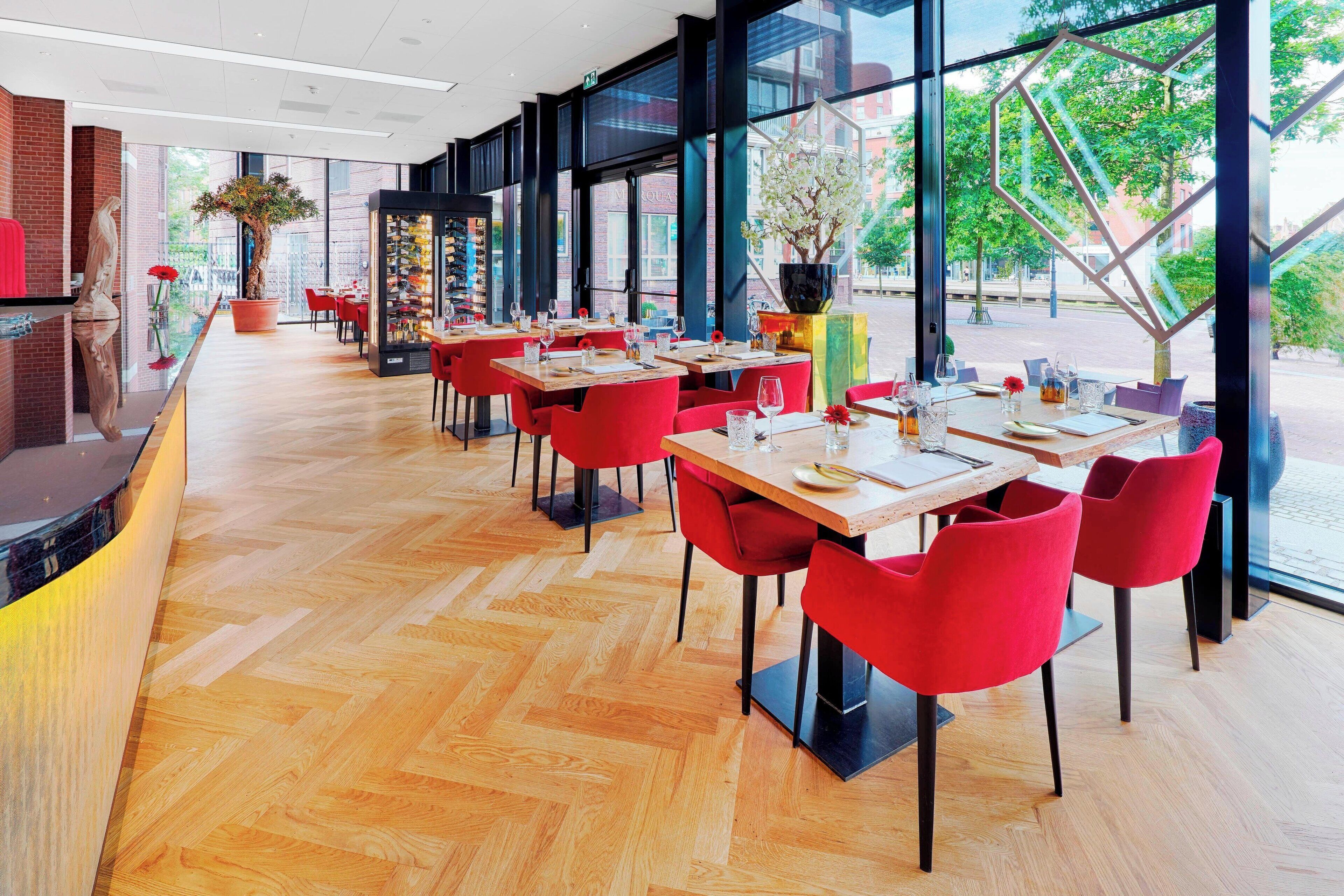 A modern restaurant with wooden flooring, red chairs, and large windows offering an outdoor view.