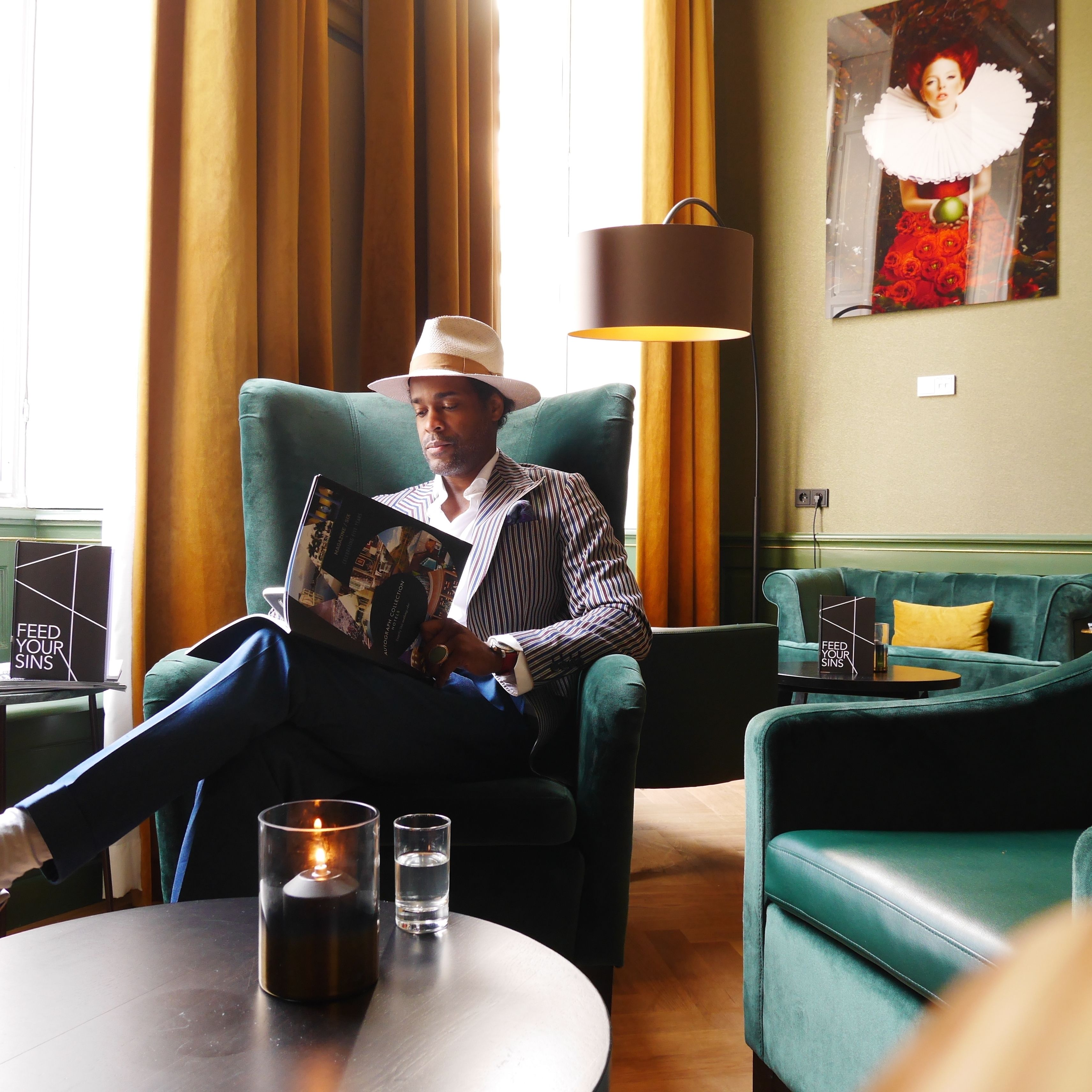 A man reading a book in a lounge area with a green sofa, a table with a candle and a glass, and a lamp.