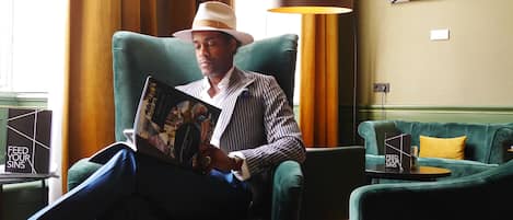 Un homme est assis dans un coin salon, en train de lire un livre. On y voit un canapé vert, une table avec une bougie et un verre, et une lampe.