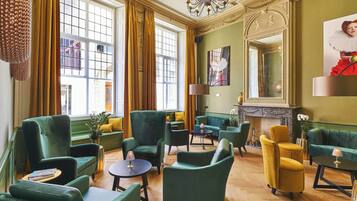 A spacious lounge with green and yellow upholstered seating, a fireplace, and large windows with curtains.