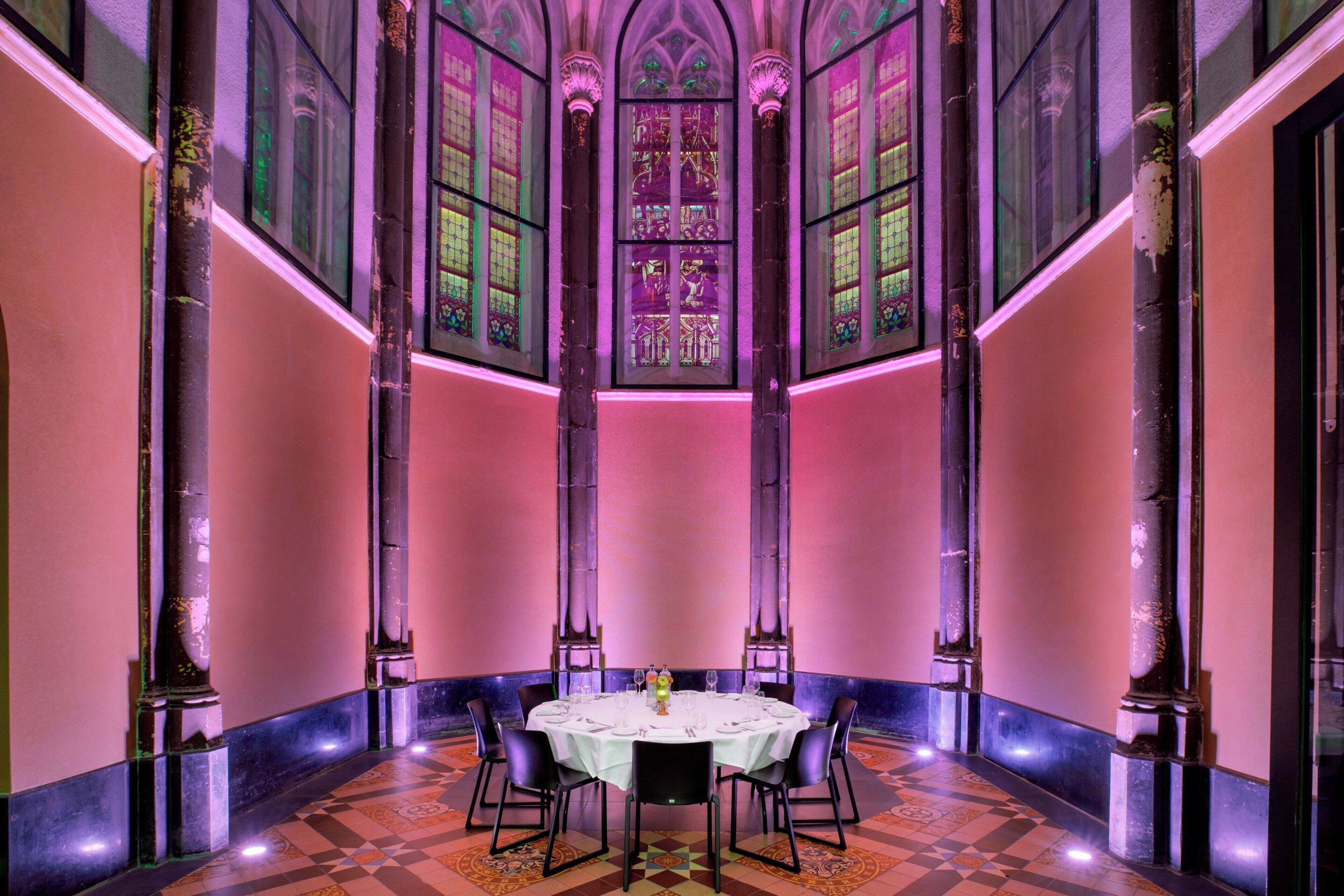 A dining area with a round table set for six, flanked by tall columns, and large stained glass windows above.
