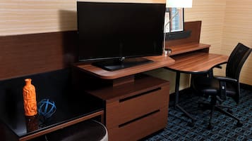 A hotel room with a flat-screen TV on a wooden desk, a black chair, a small trash bin, and a lamp on a side table.