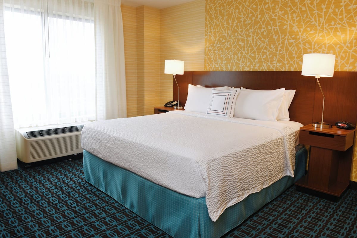Une chambre d’hôtel avec un grand lit, deux tables de chevet, une petite table, une lampe fixée au mur et une fenêtre avec des rideaux.