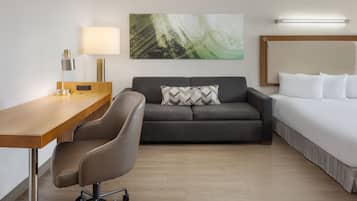 A modern hotel room with a wooden desk, a gray sofa, a bed with white linens, and a large abstract painting on the wall.