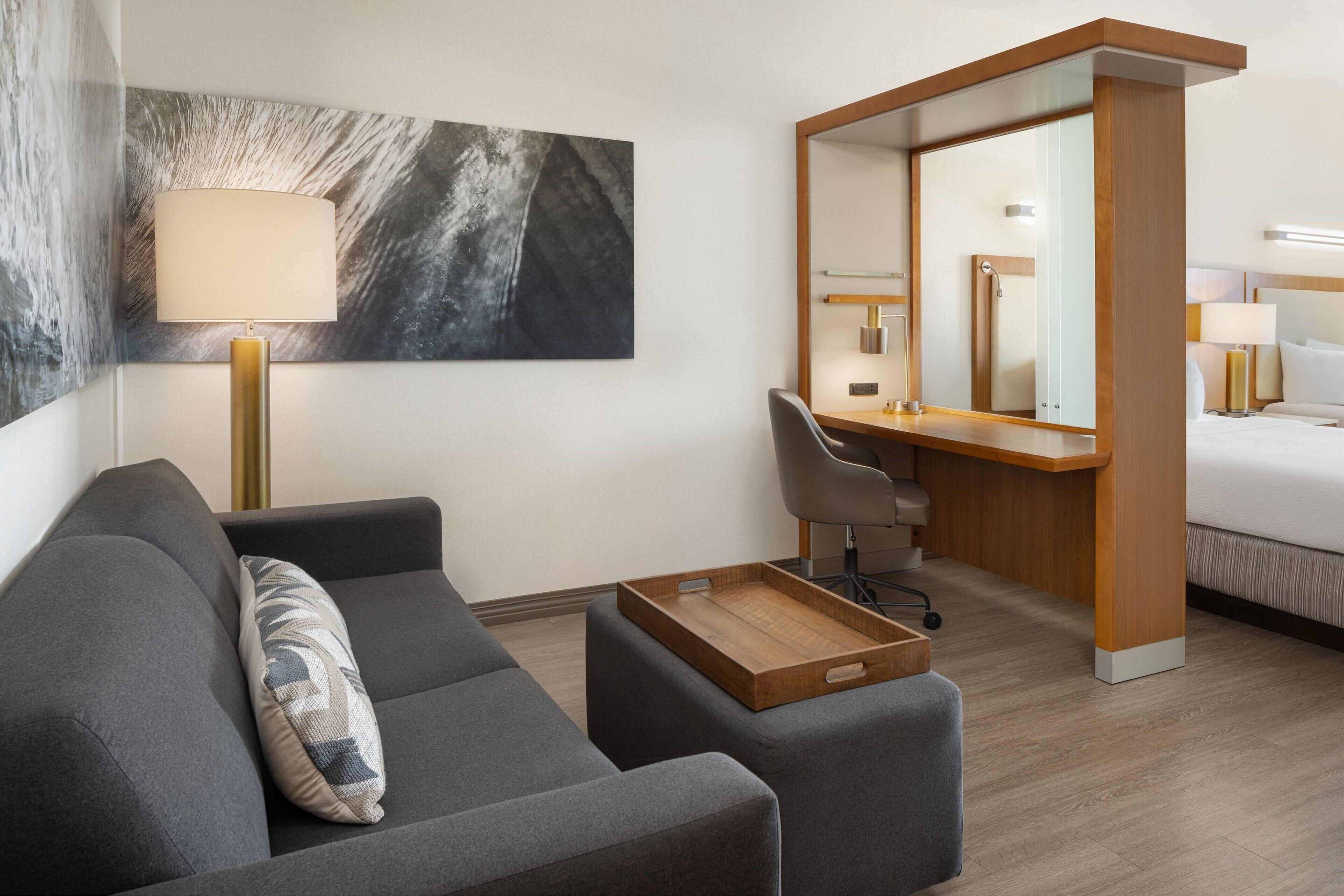 A modern hotel room with a grey sofa, a wooden side table, a desk with a chair, a bed, a large mirror, and a wall-mounted lamp.