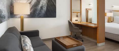 Une chambre d’hôtel moderne avec un canapé gris, une table d’appoint en bois, un bureau avec une chaise, un lit, un grand miroir et une lampe fixée au mur.