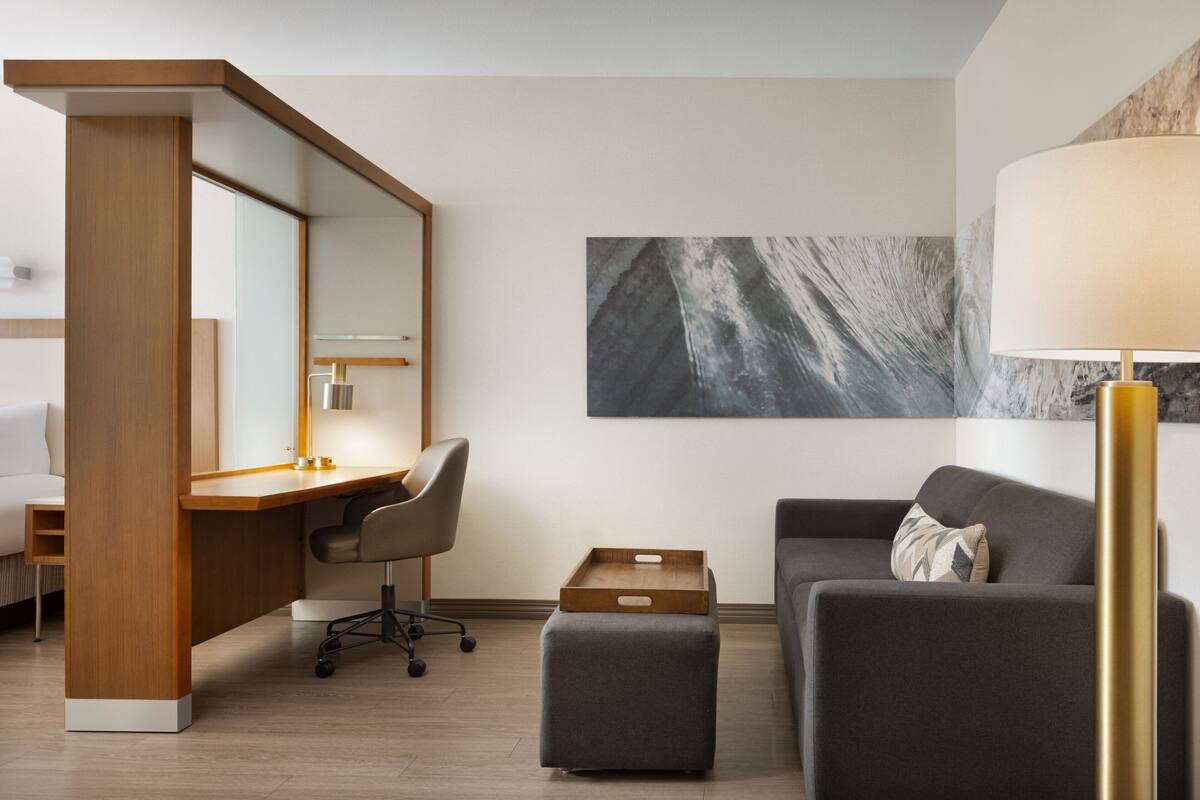 A modern hotel room with a wooden desk, a gray sofa, a small ottoman, a large mirror, and a wall-mounted lamp.