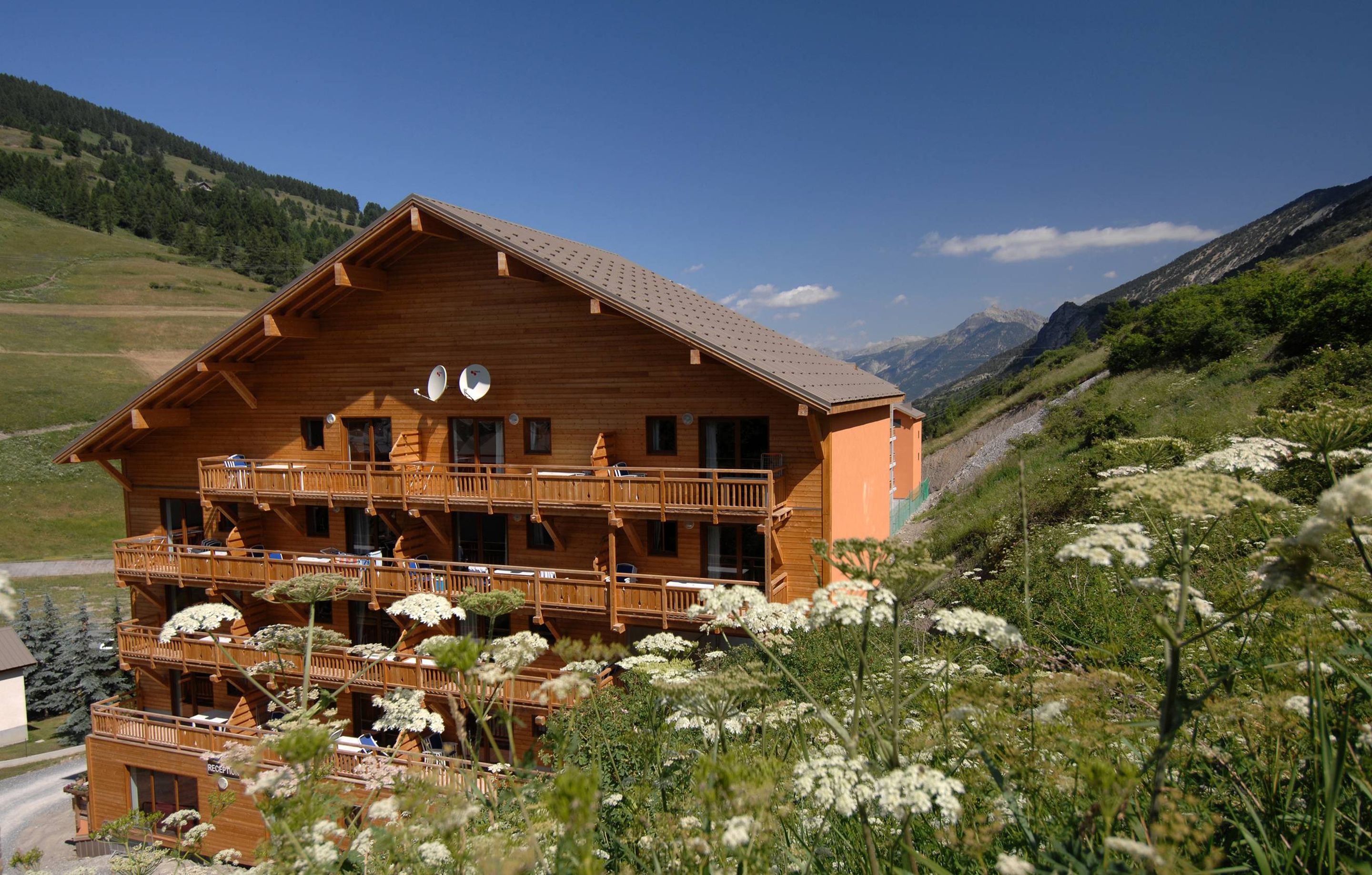 Un chalet en bois doté de nombreux balcons, entouré d’un paysage montagneux et d’une végétation luxuriante.