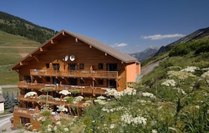A wooden chalet with multiple balconies, surrounded by a mountainous landscape and lush greenery.