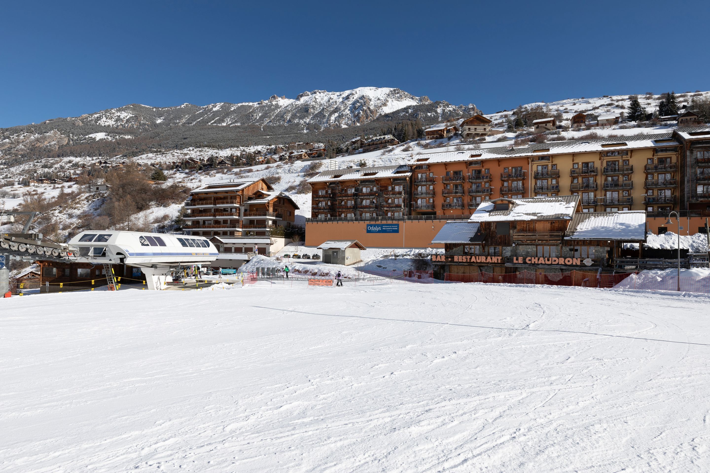 Ein schneebedecktes Skigebiet mit Gebäuden, darunter ein Restaurant namens "Le Chaudron", und einer in Betrieb befindlichen Seilbahn.