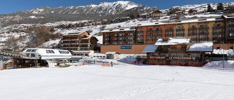 Ein schneebedecktes Skigebiet mit Gebäuden, darunter ein Restaurant namens "Le Chaudron", und einer in Betrieb befindlichen Seilbahn.
