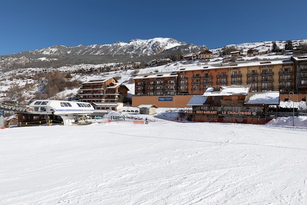 A snow-covered ski resort with buildings, including a restaurant named "Le Chaudron," and a ski lift in operation.