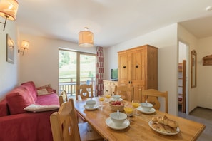 A dining area with a wooden table set for breakfast, including croissants, fruit, and orange juice. There is a red sofa and wooden cabinets in the background. - Résidence Odalys Pra Sainte Marie (Vars)